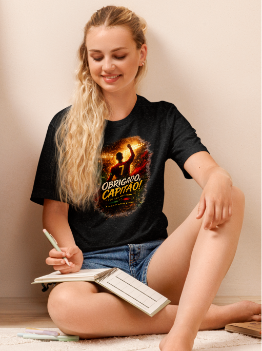 A woman sits on a light-colored floor next to a wooden dresser, wearing a black graphic t-shirt with a fiery orange and yellow design that reads 'OBRIGADO, CAPITAO!'. A matching poster is displayed on top of the dresser, and a green houseplant is on the left side of the scene.