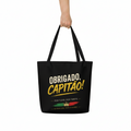A black canvas tote bag with black handles, held by a hand against a plain white background. The bag features a graphic print with the text 'OBRIGADO, CAPITAO!' in white and yellow, along with a small Portuguese flag and crown design.
