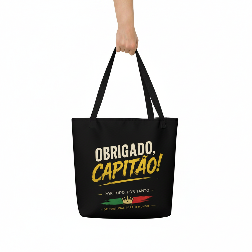 A black canvas tote bag with black handles, held by a hand against a plain white background. The bag features a graphic print with the text 'OBRIGADO, CAPITAO!' in white and yellow, along with a small Portuguese flag and crown design.
