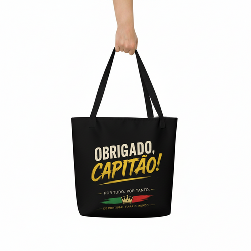 A black canvas tote bag with black handles, held by a hand against a plain white background. The bag features a graphic print with the text 'OBRIGADO, CAPITAO!' in white and yellow, along with a small Portuguese flag and crown design.